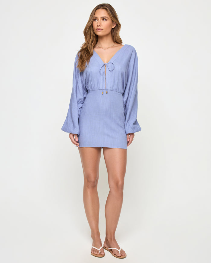 Front view of a model wearing the LSPACE Deia Mini Dress in chambray, featuring a V-neckline with tie-front closure and gold hardware, wide balloon sleeves, elastic waistband, and mini length in chambray blue linen fabric, styled with white thong sandals.