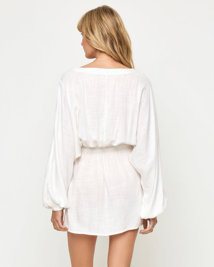 Back view of a model wearing the LSPACE Deia Mini Dress in cream, showing the open V-back, wide balloon sleeves, elastic waistband, and mini length in cream linen fabric.