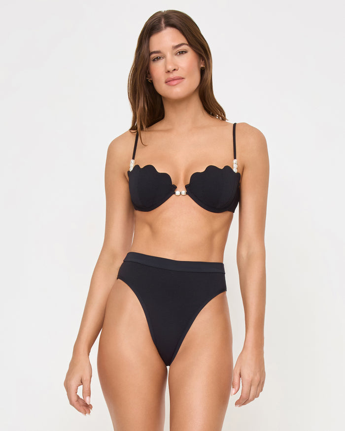Front view of a model wearing the LSPACE black Frenchi Bikini Bottom with a high-waisted style and high-cut leg, paired with a black scalloped Sunrise Shell Bikini Top.