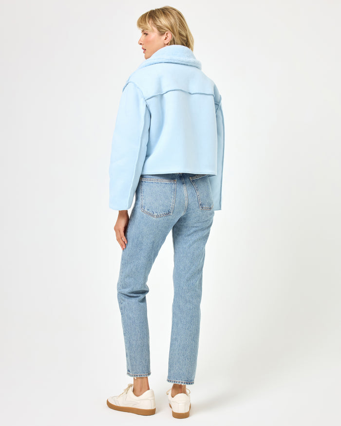Back view of a model wearing the LSPACE Kalina Jacket in ice blue, showing the smooth fleece reverse side and wide collar of the cropped reversible jacket, styled with jeans and white sneakers.