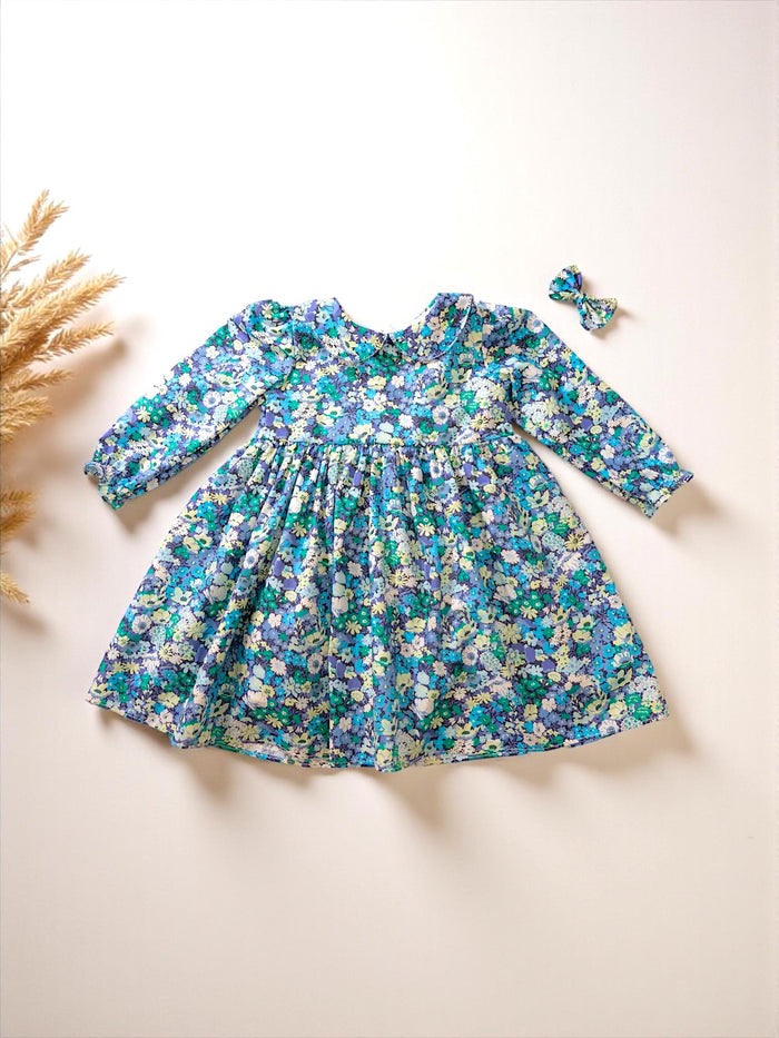 May include: A blue floral print dress with long sleeves and a Peter Pan collar. The dress features a full skirt and is paired with a matching bow. The dress is decorated with a pattern of small flowers in shades of blue, green, and white.