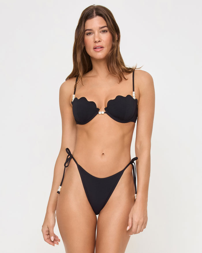 Straight-on front view of a model wearing the black LSPACE Lucia Bikini Bottom with pearl side ties and a Sunrise Shell Bikini Top in a studio setting.