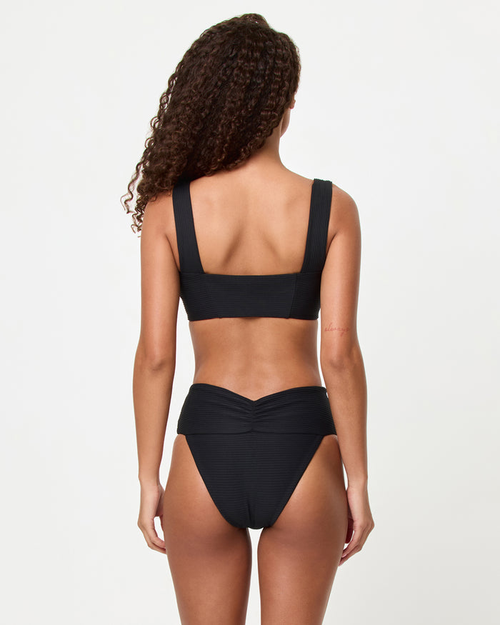 Back view of a model wearing the LSPACE black Bardot Bikini Bottom, a ribbed high-waist style showing the gathered center back waistband detail, paired with the black Marlee Bikini Top.