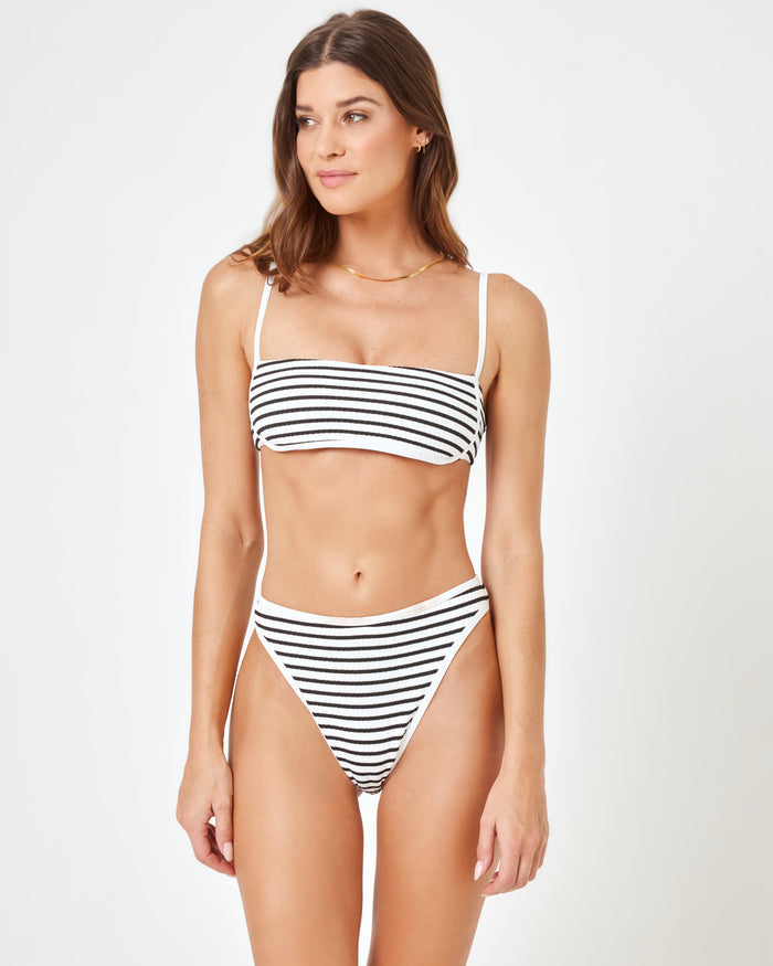 Front view of a model in a studio wearing the LSPACE Sail Along Stripe Nora Bikini Bottom in white and black horizontal stripes with a mid-rise fit and high-cut leg openings, styled with a matching Hazel Bikini Top.