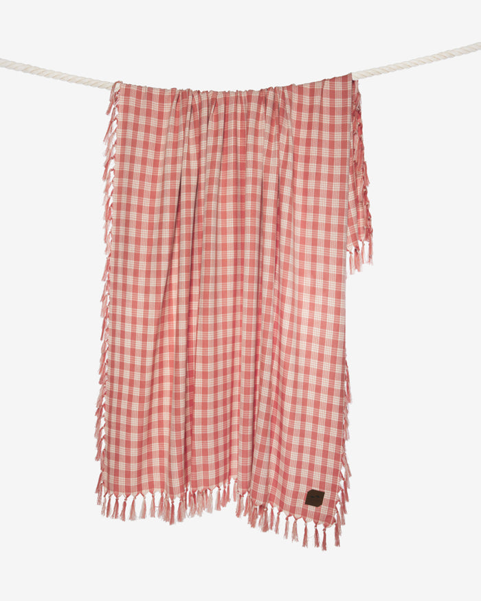 The Slowtide Palaka Lightweight Beach Blanket in guava hanging on a rope, showing the pink gingham check pattern with fringe.