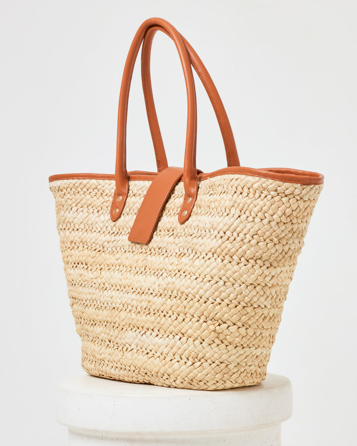 Side view of the Alfie Bag in natural, a large woven straw tote with brown leather handles, on a white pedestal.