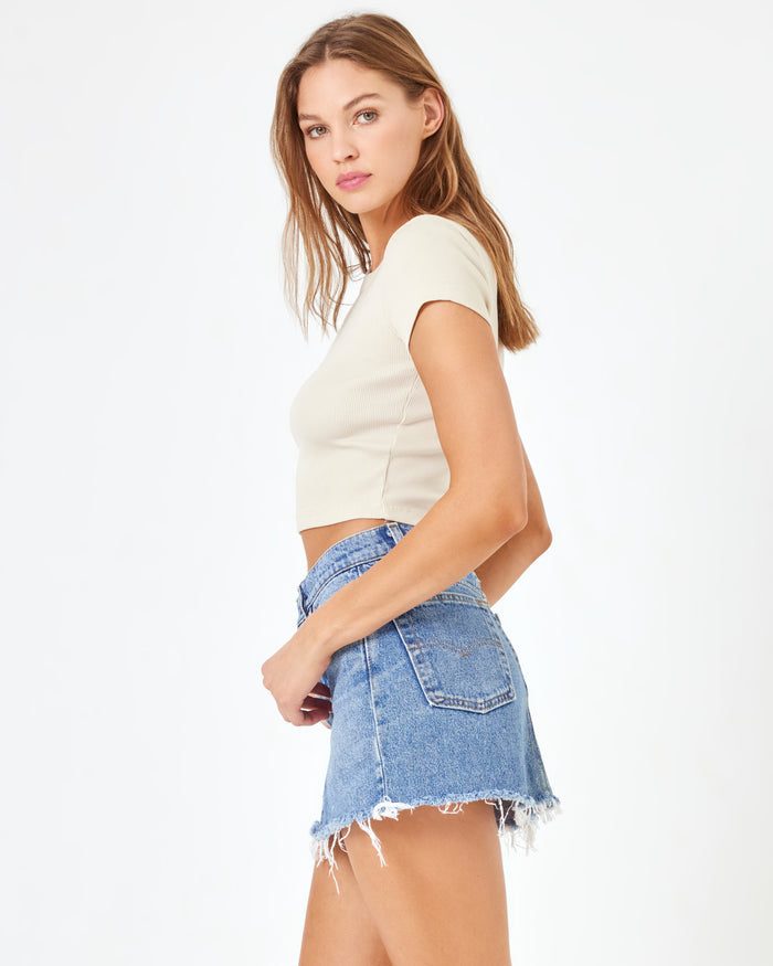 Side view of a model wearing the LSPACE aligrace Mini Skirt in light wash denim blue, a denim mini skirt with a frayed hem, styled with a cream crop tee.