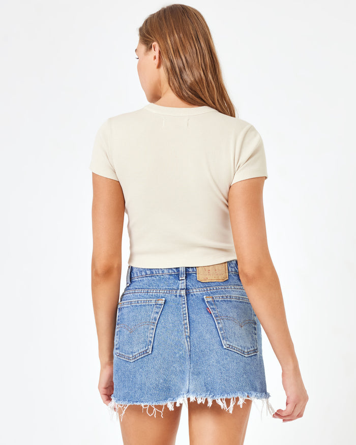 Back view of a model wearing the LSPACE aligrace Mini Skirt in light wash denim blue, a denim mini skirt with a frayed hem, styled with a cream crop tee.