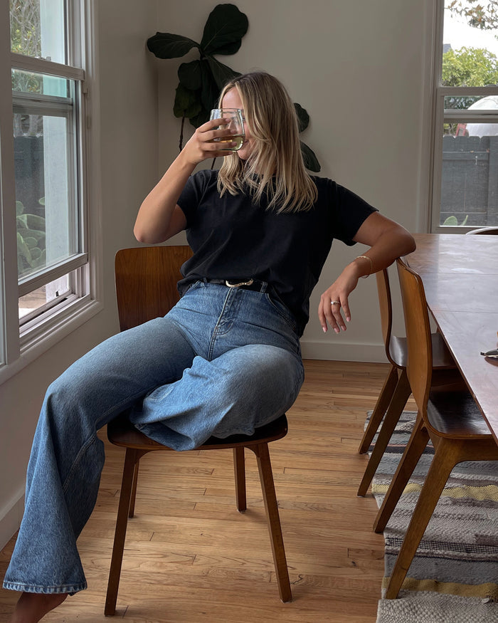 Lifestyle front view of a model seated at a dining table wearing the LSPACE All Day Top in black, styled with light wash wide-leg jeans.