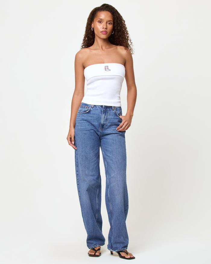 Full-length front view of a model wearing the LSPACE Andie Top in yallspace, a white ribbed strapless tube top with an embroidered cowboy boot graphic, styled with medium wash wide-leg jeans and black sandals.