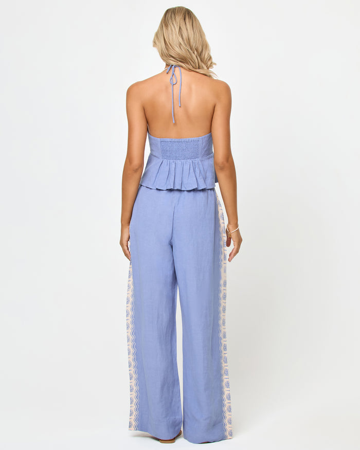 Back view of a model wearing the LSPACE April Top in shell, showing the open back with halter neck tie, smocked band, and peplum ruffle hem, paired with matching wide-leg pants.