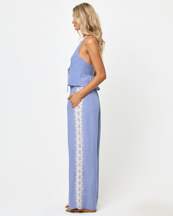 Side view of a model wearing the LSPACE April Top in shell, a periwinkle blue halter top with a peplum ruffle hem, styled with matching wide-leg pants with cream embroidered trim.