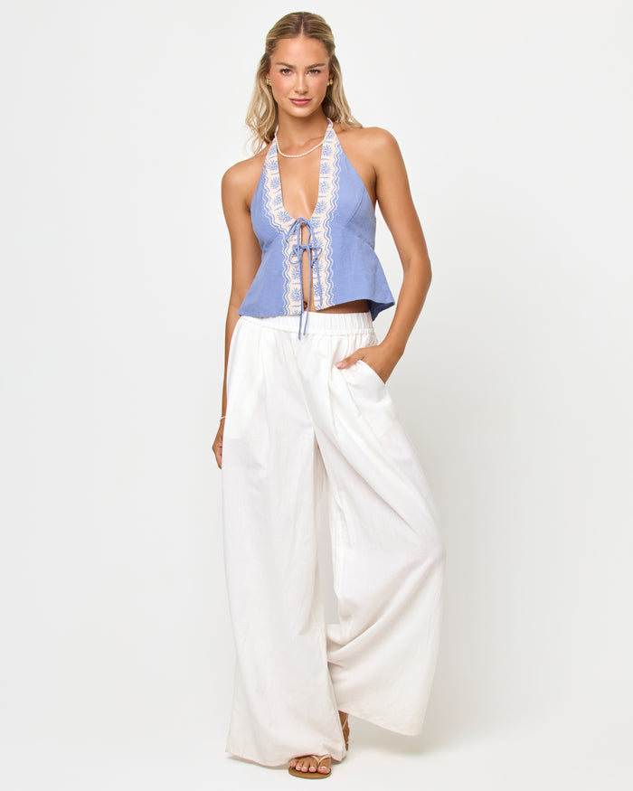 Full-length front view of a model wearing the LSPACE April Top in shell, a periwinkle blue halter top with cream embroidered trim and lace-up front closure, styled with white wide-leg pants and sandals.