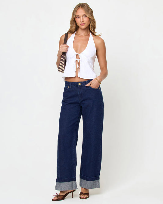 Front view of a model wearing the LSPACE April Top in white, featuring a deep V-neckline with a tie-front detail, halter neck, and peplum hem in white linen fabric, styled with dark navy wide-leg cuffed jeans and a woven shoulder bag.