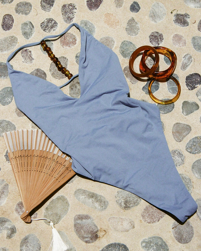 Off-model front view of the LSPACE Arden One Piece Swimsuit in chambray blue on a pebbled stone surface with accessories.