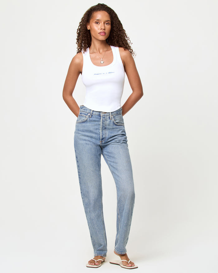 Full-length front view of a model wearing the LSPACE Ash Tank in happier in a bikini, a white cropped ribbed racerback tank with embroidered script text on the chest, styled with light wash straight-leg jeans and white sandals.