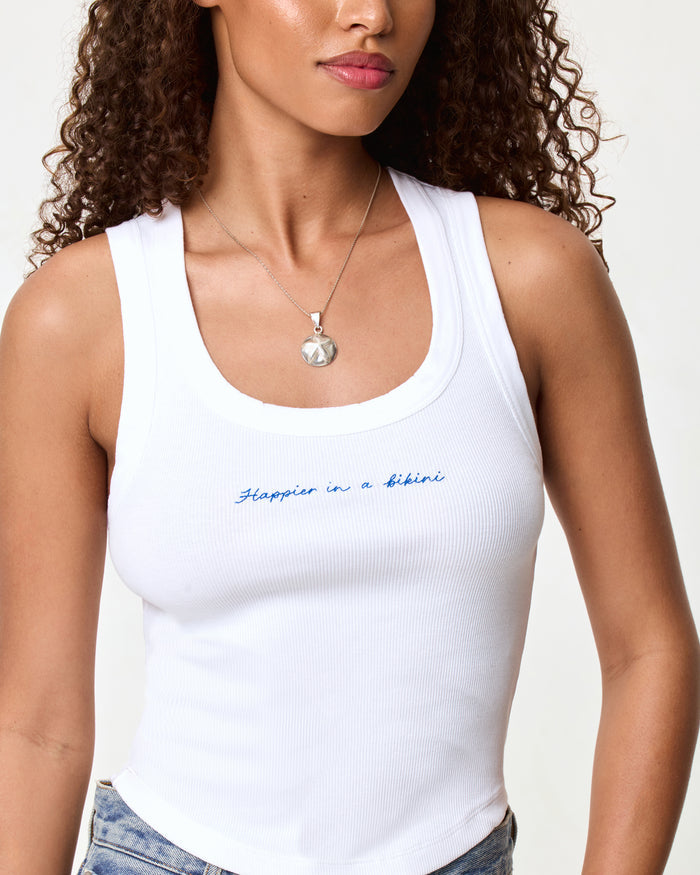 Close-up of the LSPACE Ash Tank in happier in a bikini, highlighting the blue embroidered script text reading "Happier in a bikini" on the white ribbed fabric.