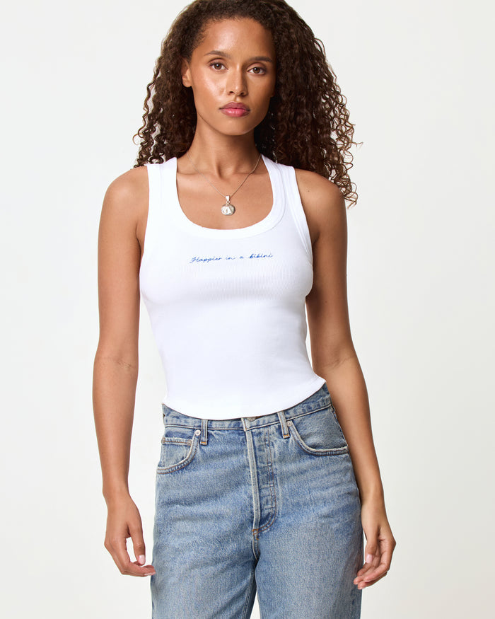 Front view of a model wearing the LSPACE Ash Tank in happier in a bikini, a white cropped ribbed racerback tank with blue embroidered script text on the chest, styled with light wash jeans.