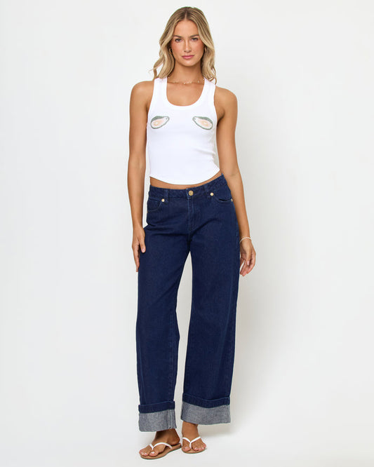 Full-length front view of a model wearing the LSPACE Ash Tank in oysters, a white cropped ribbed racerback tank with an oyster shell graphic on the chest, styled with dark navy wide-leg cuffed jeans and sandals.