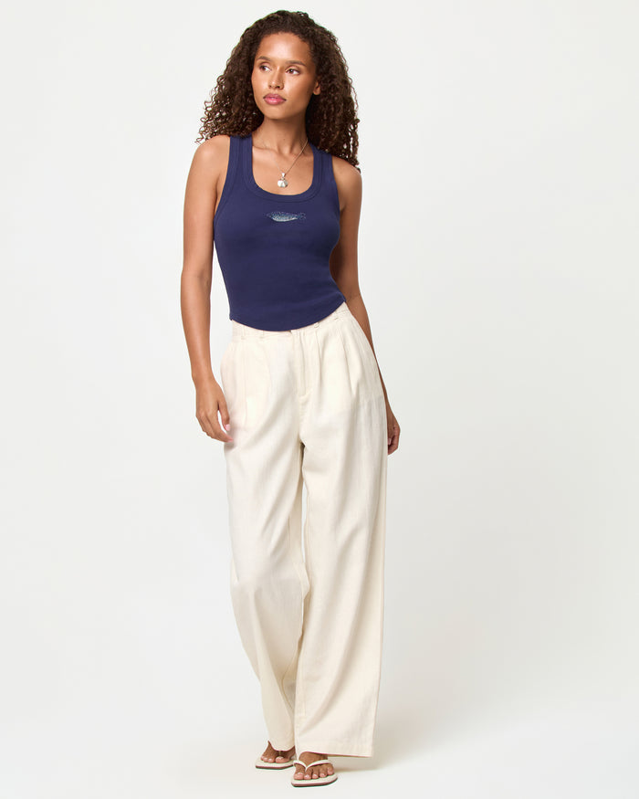 Full-length front view of a model wearing the LSPACE Ash Tank in sardine, a navy blue cropped ribbed racerback tank with a rhinestone sardine fish graphic, styled with cream wide-leg pants and flip-flop sandals.