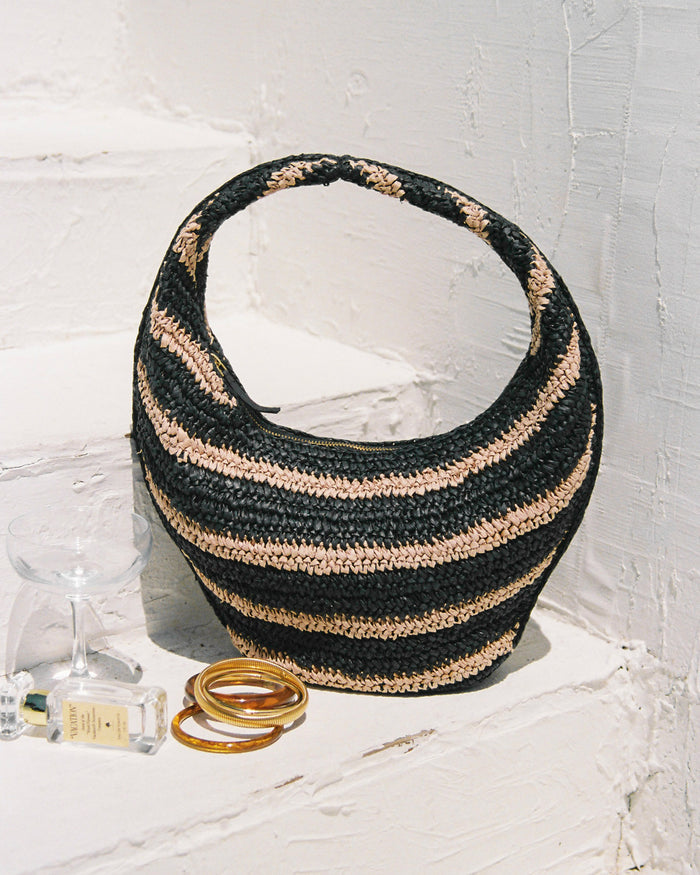 Lifestyle image of the Aya Bag in natural-black, a crescent-shaped woven raffia hobo bag with black and natural horizontal stripes, styled on a white shelf with bangles and a perfume bottle.