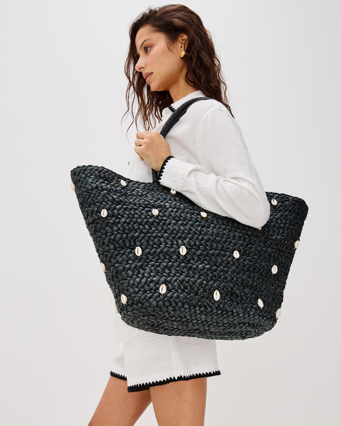 Model carrying the Baltic Seas Tote in black over the shoulder, a large woven raffia tote bag with scattered cowrie shell embellishments, paired with a white button-down shirt and white skirt.