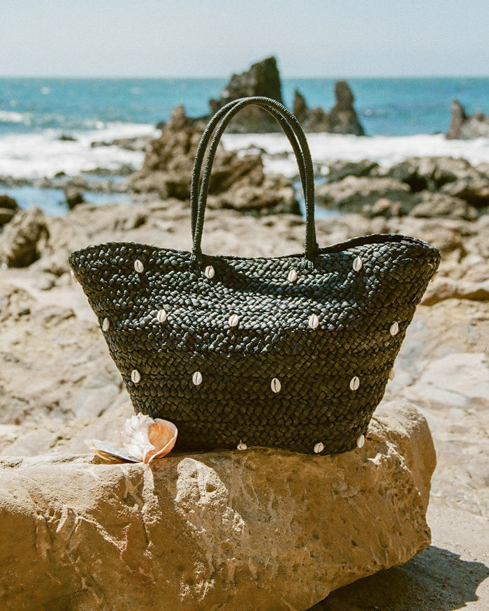 Lifestyle image of the Baltic Seas Tote in black resting on a rocky coastal ledge with a seashell beside it and the ocean in the background.