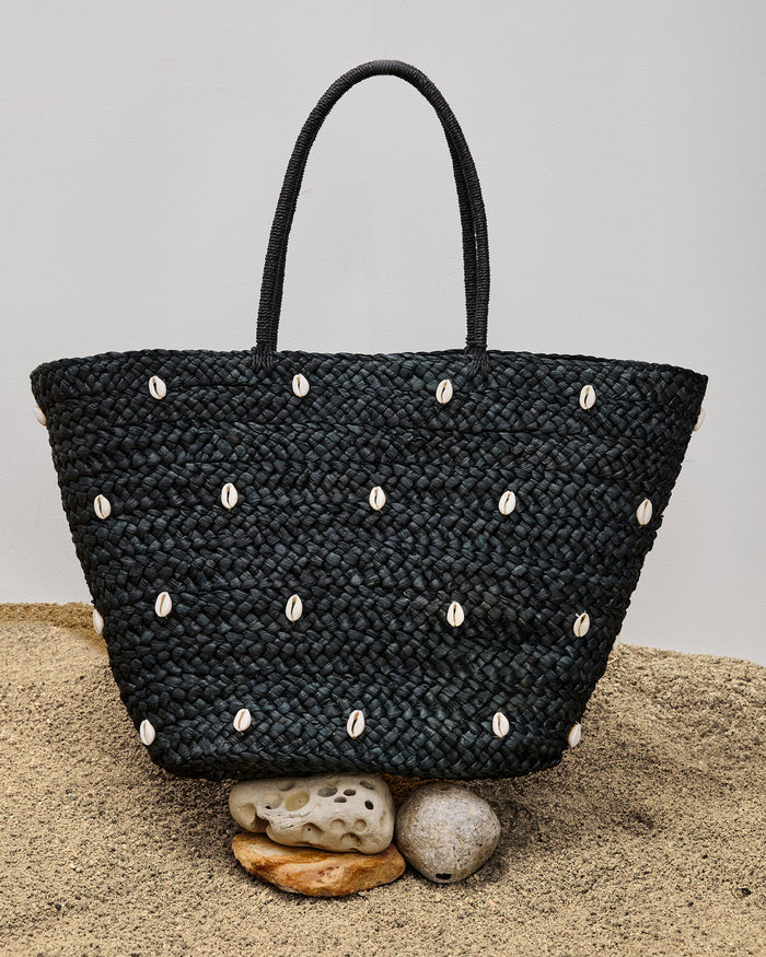 Off-model front view of the Baltic Seas Tote in black with cowrie shell embellishments, displayed on sand with sea stones in front.