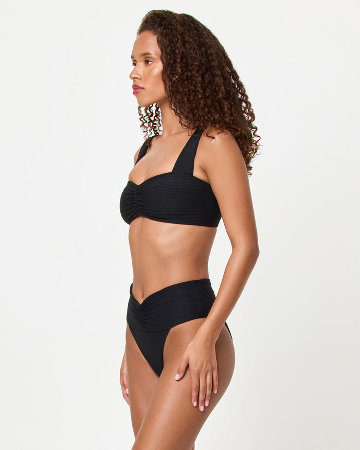 Side view of a model wearing the LSPACE black Bardot Bikini Bottom, a ribbed high-waist style with gathered waistband detail, paired with the black Marlee Bikini Top.