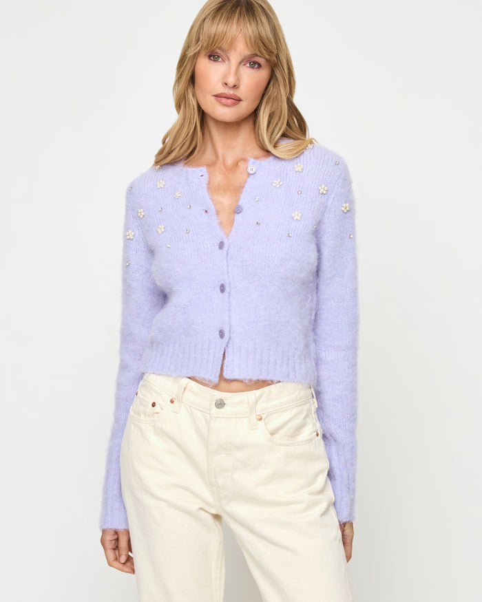 Close-up of a model wearing the LSPACE Brynn Cardigan in lavender ice, featuring a cropped fuzzy knit silhouette in pale lavender with pearl floral embellishments and a button-front V-neckline, styled with cream wide-leg jeans.