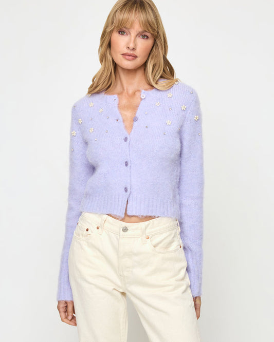 Close-up of a model wearing the LSPACE Brynn Cardigan in lavender ice, featuring a cropped fuzzy knit silhouette in pale lavender with pearl floral embellishments and a button-front V-neckline, styled with cream wide-leg jeans.