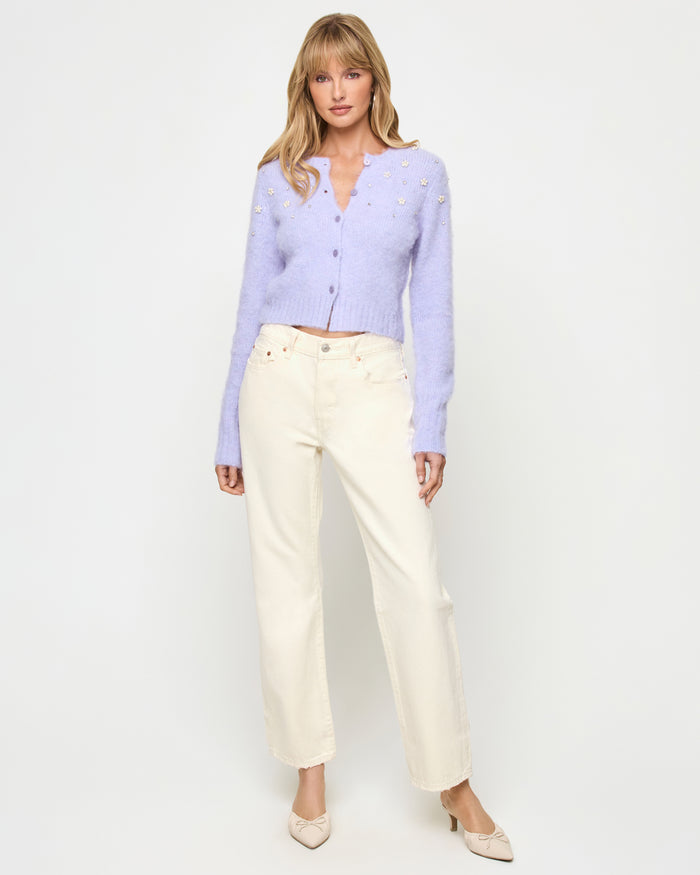 Front view of a model wearing the LSPACE Brynn Cardigan in lavender ice, featuring a cropped fuzzy knit silhouette in pale lavender with pearl floral embellishments and button-front V-neckline, styled with cream wide-leg jeans and bow-toe heels.