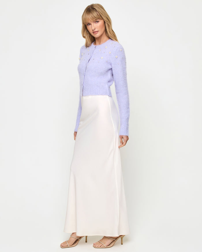 Side view of a model wearing the LSPACE Brynn Cardigan in lavender ice, showing the cropped fuzzy knit profile with long sleeves and pearl floral embellishments, styled with a cream satin maxi skirt.