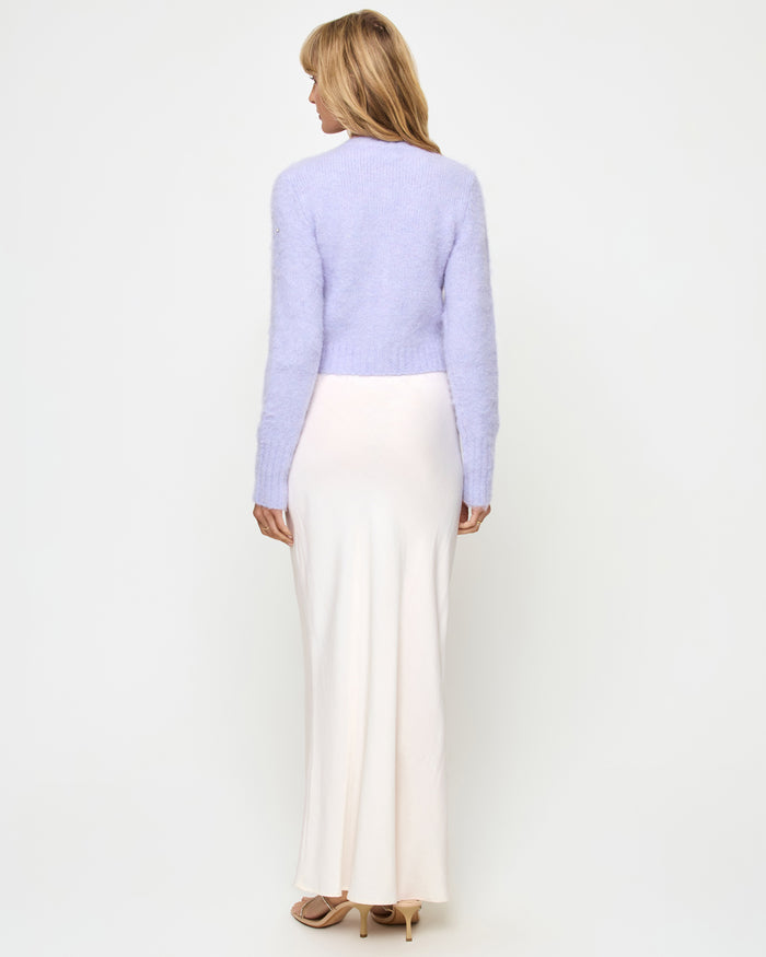 Back view of a model wearing the LSPACE Brynn Cardigan in lavender ice, showing the cropped fuzzy knit silhouette in pale lavender, styled with a cream satin maxi skirt.