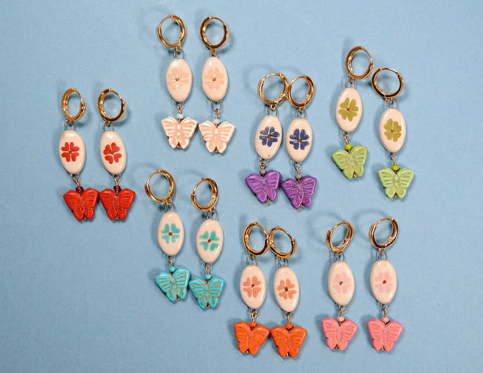 Six pairs of handmade dangle earrings arranged on a light blue background. Each pair features a gold-tone hoop and a dangling oval charm decorated with a small flower, followed by either a butterfly or clover charm in various pastel and bright colors including red, orange, pink, purple, green, and blue.