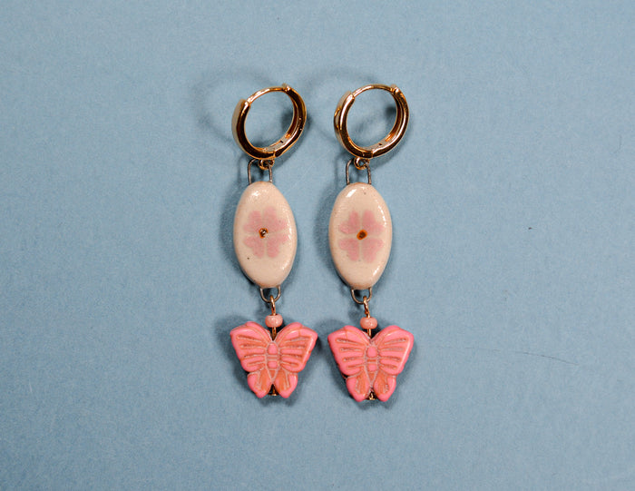 A pair of dangling earrings with round gold-tone hoops, oval pale pink stone-like centers, and pink butterfly charms below, displayed on a light blue background.