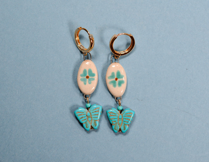 A pair of handmade drop earrings featuring gold-tone hoops, light pink oval beads with turquoise floral designs, and turquoise butterfly charms with gold outlines, displayed on a light blue background.