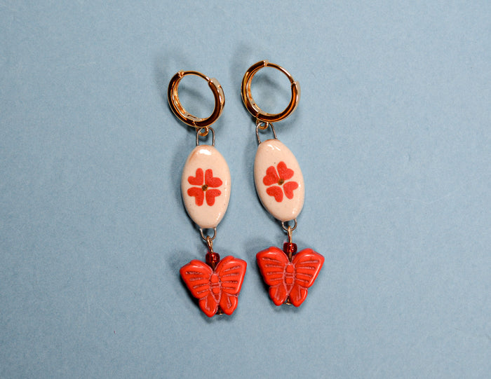 A pair of handmade dangle earrings featuring gold hoop backs, white oval beads with red flower motifs, and red butterfly charms, displayed on a light blue background.