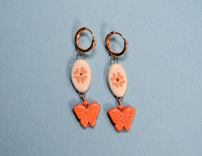 A pair of retro-style drop earrings featuring gold hoop backs, white oval beads with pink floral engravings, and dangling orange butterfly charms, set against a solid light blue background.