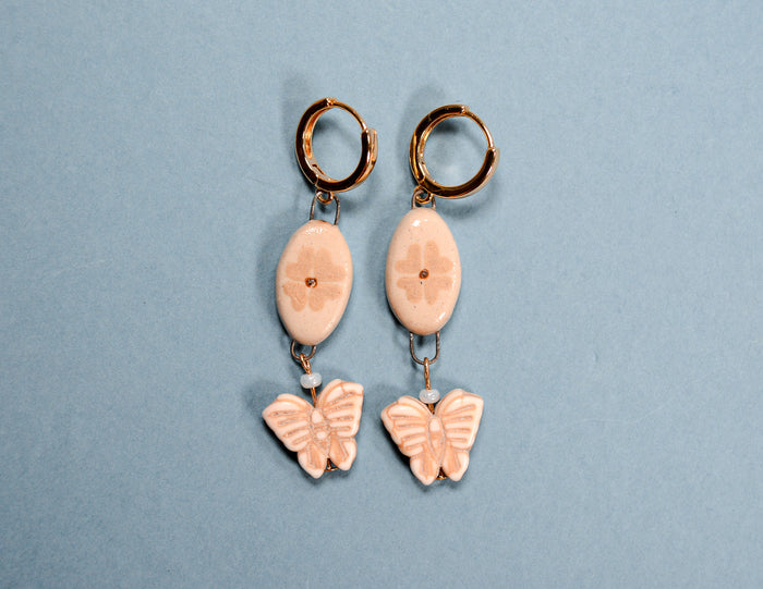 A pair of dangling earrings with gold-tone hoops, pale pink oval beads with floral details, and butterfly charms, displayed on a light blue background.