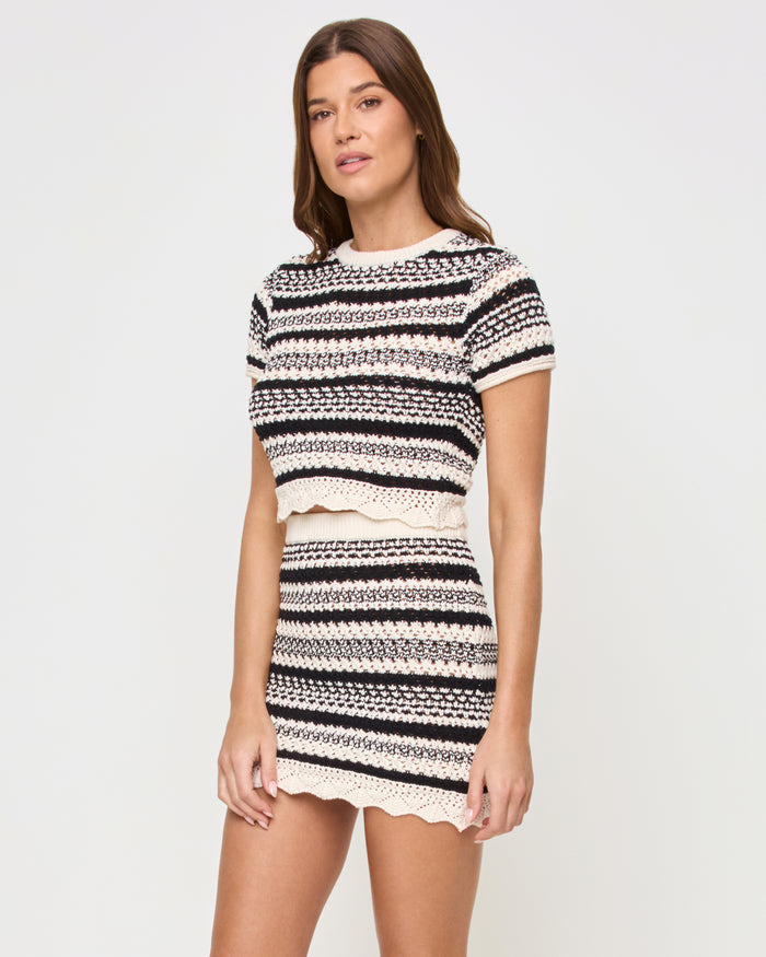 Three-quarter front view of a model wearing the LSPACE Caicos Skirt in caicos stripe, a black-and-cream crochet mini skirt with horizontal stripes and scalloped hem, styled with the matching Caicos Top.