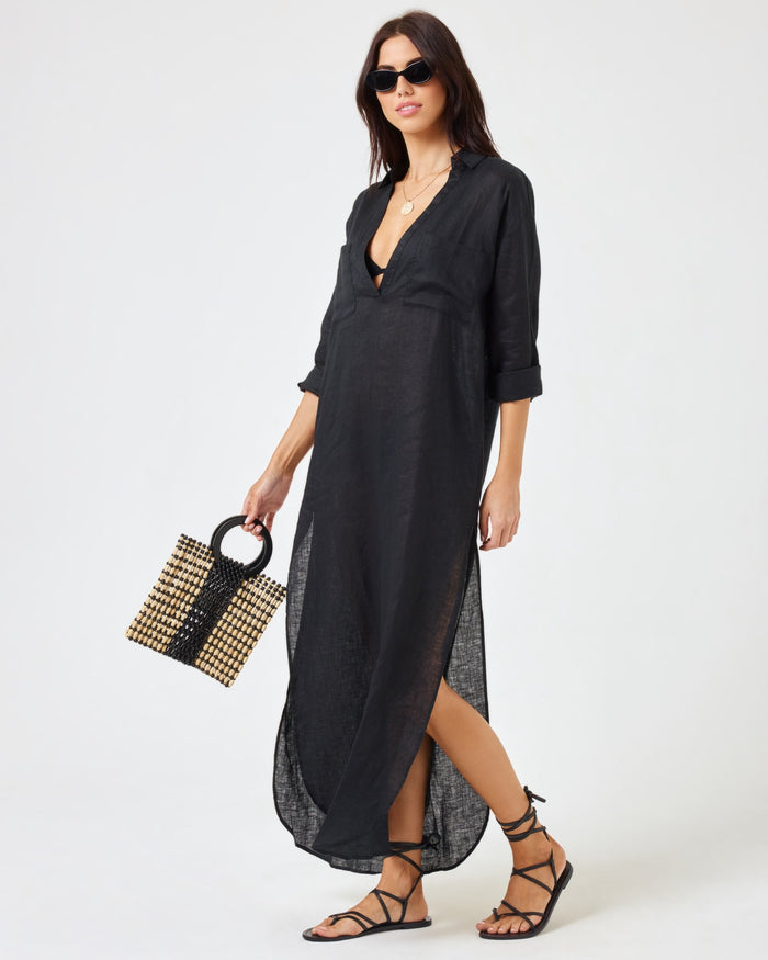 Angled front view of a model wearing the LSPACE Capistrano Tunic in black, a sheer linen maxi with a deep V-neck collar and side slit, styled with sunglasses and a woven bag.