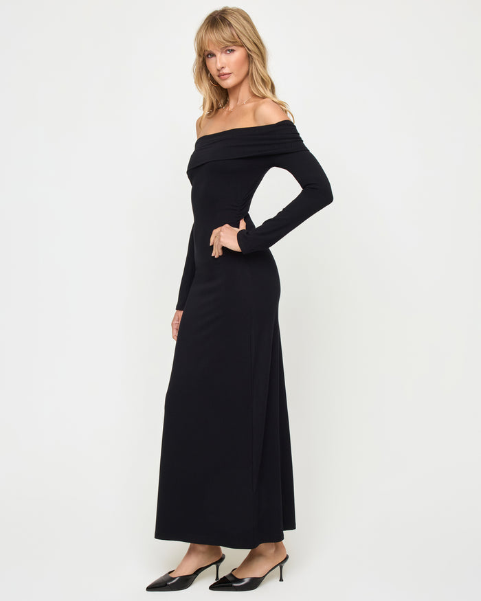 Side view of a model wearing the LSPACE Carrie Dress in black, showing the off-the-shoulder silhouette with long sleeves and body-hugging maxi length in ribbed black knit fabric.