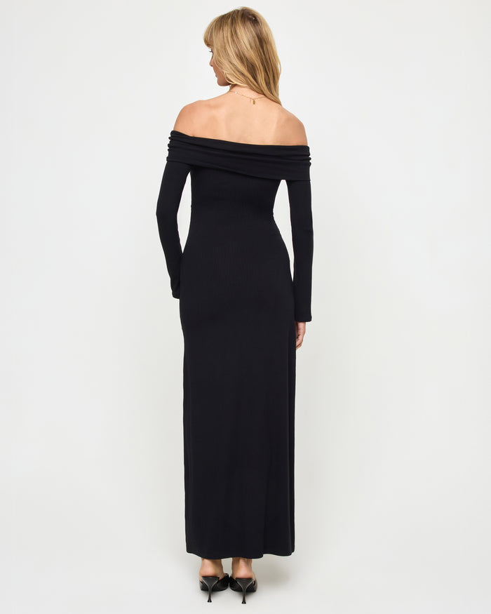 Back view of a model wearing the LSPACE Carrie Dress in black, showing the off-the-shoulder folded neckline band, long sleeves, and body-hugging maxi silhouette in ribbed black knit fabric.