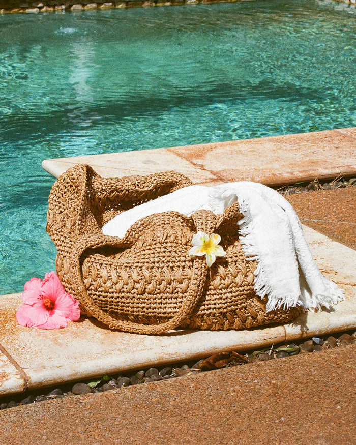 Lifestyle image of the Changing Tides Bag in natural resting by a pool with a white towel draped over it and tropical flowers nearby.
