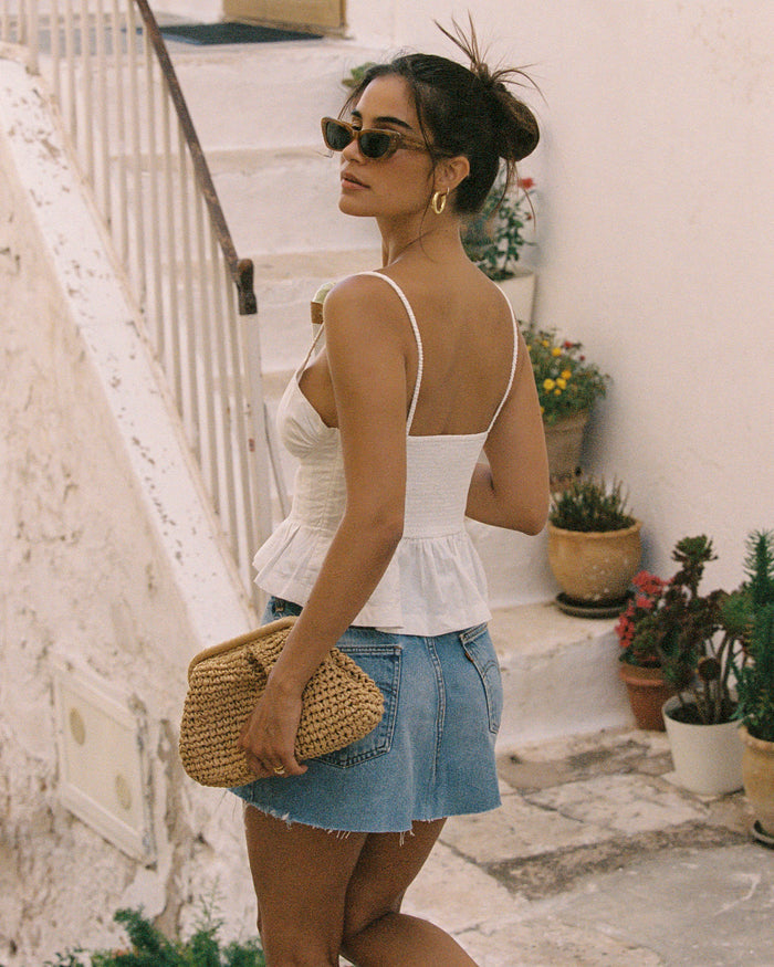 Back view of a model wearing the LSPACE Clover Top in white, showing the open square back with spaghetti straps and pleated peplum hem in white linen fabric, styled with a denim mini skirt, photographed on outdoor stone steps.