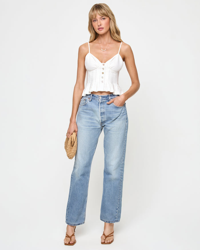 Front view of a model wearing the LSPACE Clover Top in white, featuring the V-neckline with spaghetti straps, button-front closure, and pleated peplum hem, styled with light-wash wide-leg jeans, a woven bag, and tan thong sandals.