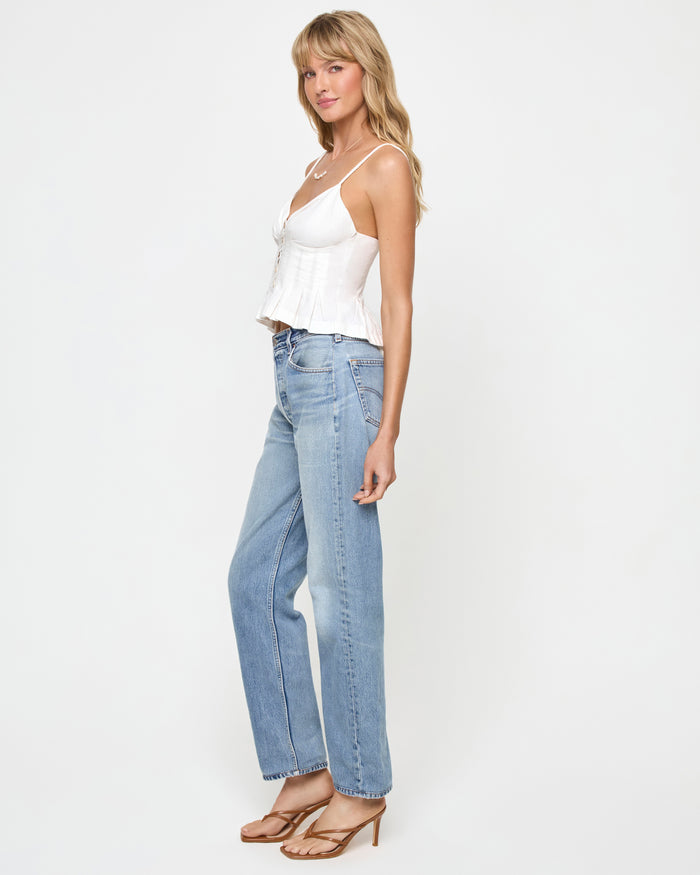 Side view of a model wearing the LSPACE Clover Top in white, showing the spaghetti-strap silhouette with button-front bodice and pleated peplum hem, styled with light-wash straight jeans and tan heeled sandals.