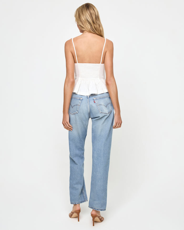Back view of a model wearing the LSPACE Clover Top in white, showing the open square back with spaghetti straps and pleated peplum hem in white linen fabric, styled with light-wash wide-leg jeans.