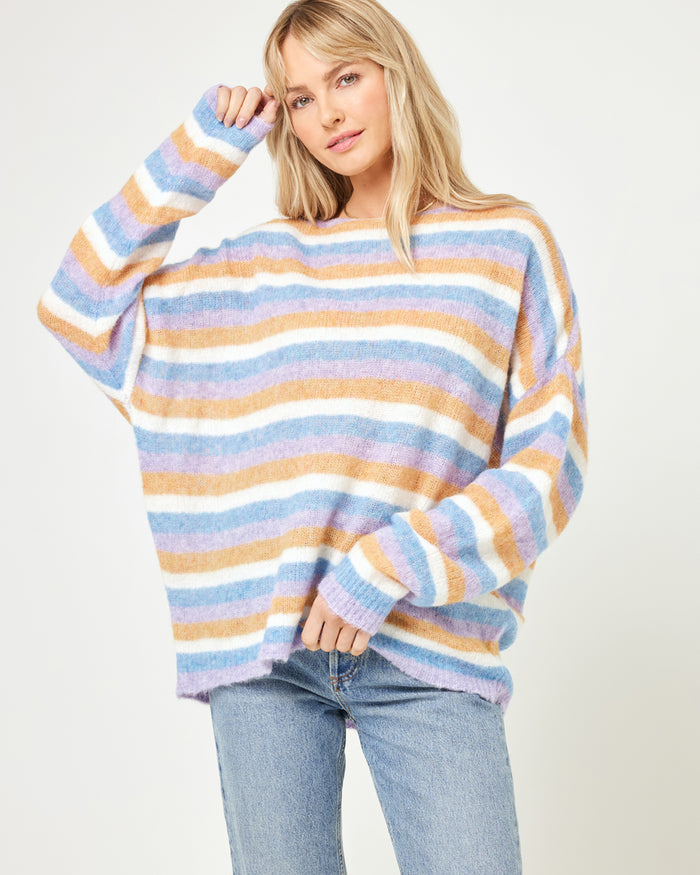Cropped front view of a model wearing the LSPACE Cozy Clouds Sweater in beautiful skies stripe, an oversized crew-neck sweater in a soft fuzzy knit with multicolor horizontal stripes in blue, tan, pink, and white, paired with jeans.